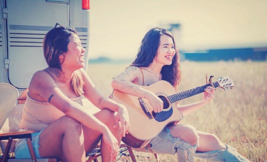 Asian chinese lesbian and her friend sit on camping chair playing guitar enjoying outdoor camping beside motor home